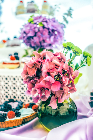 flower bouqet of  green chrysanthemum and  pink hydrangea on the violet tablecloth and white backgroun with the soft focused assorted tarts and cupcakes with berriesの写真素材