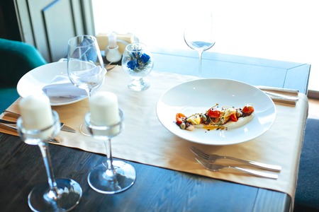 Dish in the white plate on the table with flower decor,candles, wineglasses and bright backgroundの写真素材