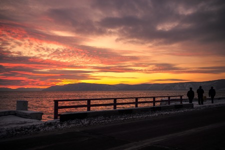 People watching sunset at embankment. Beautiful yellow-pink cover with cloudy sky and mountains.の写真素材