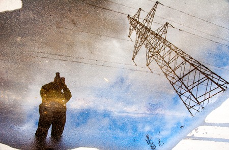 Photographer reflection on a water puddle with blue sky and electric tower