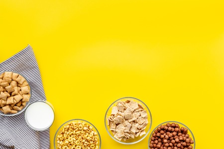 Various corn cereals in bowls and milk on yellow background top view copyspace