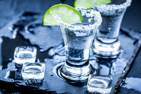 Silver tequila shots with ice cubes and fresh lime slices on black table backgroundの写真素材