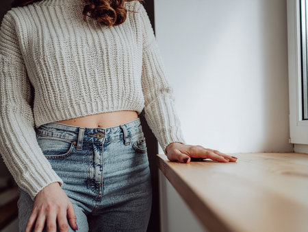 Unrecognizable young woman in jeans and white sweater standing near the window.の素材