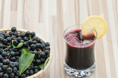 Fresh juice of chokeberry or Aronia melanocarpa in glass with ice and lemone slice, berry in basket on wooden backgroundの写真素材