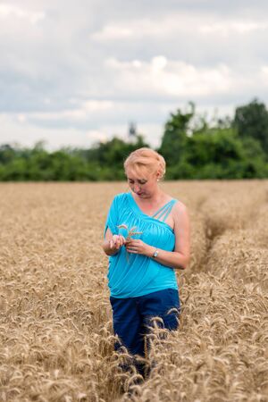Beautiful woman stand in golden wheat field and hold ear of wheat, with cloudy blue sky background. Liberty, happy summer, peace of mind or agriculture concept.の写真素材