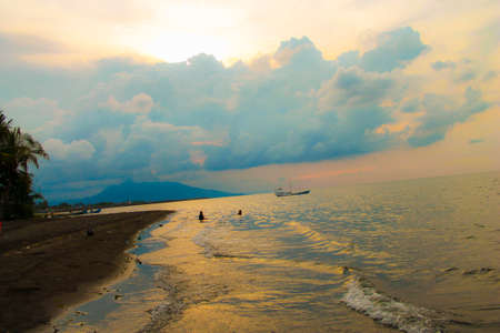 Indonesia beach with beautiful sunset tropical landscape, there are a sailing boat, mountain, waves, sand, trees, and clody sky.の写真素材