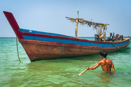 A fisherman wades out in waist deep water to his colourful wooden boat off the beach at Ngapali, Myanmar, against a clear blue skyの写真素材