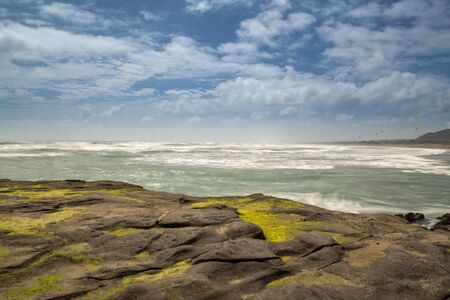 The Pacific Ocean at Muriwai Beach on the West coast of New Zealand, shot across the algae covered rocks with a long exposure to smooth out the water and give some motion to the clouds in blue skyの写真素材