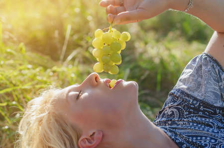 Young beautiful blonde woman eating bunch of grapes in nature in sun rays.の写真素材