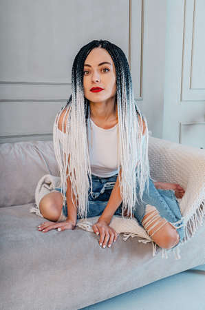 Sexy woman with dreadlocks. White t-shirt and blue jeans. Sitting on gray sofa on knees. Hands...