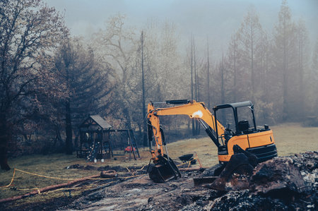 Yellow small excavator clears the area after a fire in the fog in the eveningの写真素材