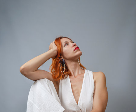 Portrait Sexy red-haired woman in white. Massive jewelry on ears. red lipstick. The head is tilted to the side. deep neckline.の写真素材