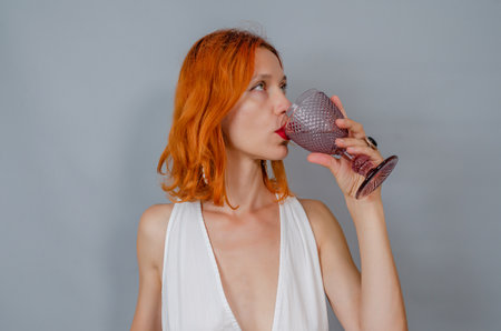 Portrait of red-haired woman in white with glass of wine. A woman drinks from glass. side view.の写真素材