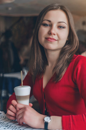Cute brunette girl in red blouse is sitting at table in a cafeの写真素材