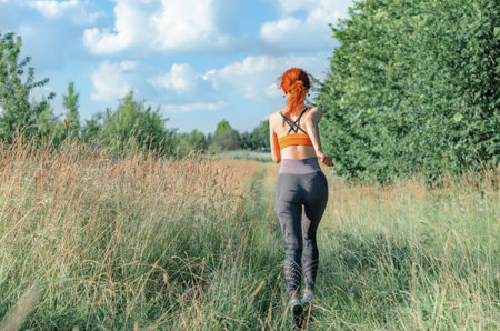 A young female runner runs along the road overgrown with grass. rear viewの写真素材