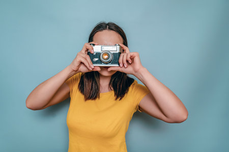A woman takes pictures with an old film camera. Yellow boot cap, black hair, blue backgroundの写真素材