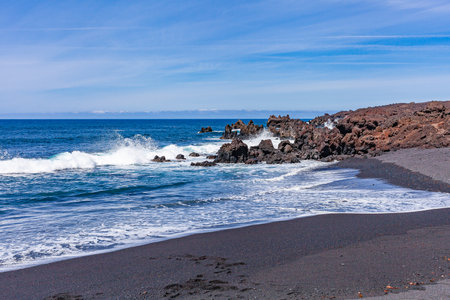 Bright contrast of white sea foam of the waves rolling onto shore with the sand and black solidified lava.の写真素材