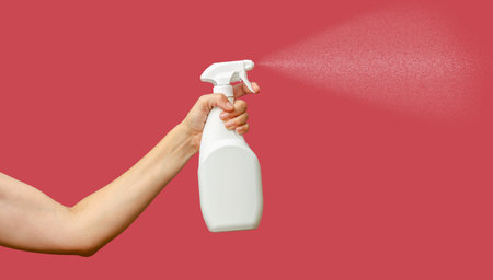 Woman's hand holds white unlabeled spray bottle with detergent. Professional cleaning. Pink backdrop