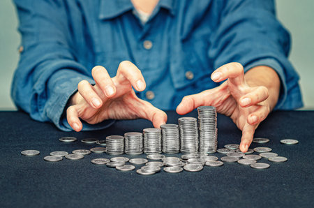 Greedy hands of woman in blue reaching for columns of coins. Concept of theft, fraud and greedの写真素材