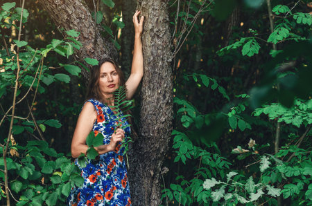 Portrait of dreamy woman near tree, looking into camera, dark forest. Hold green fern in her handの写真素材