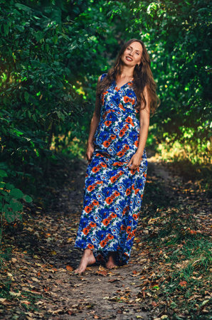 Joyful woman with long hair walks barefoot along forest path. She holds long dress in hands and laughsの写真素材