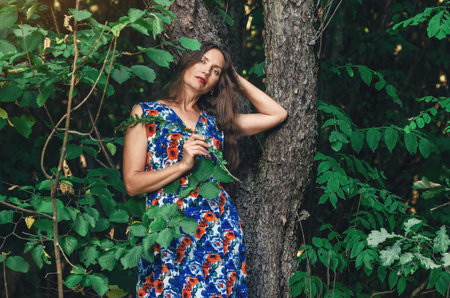 Portrait of dreamy woman near tree, look into camera, tilting head to side. Hold green fern in handの写真素材