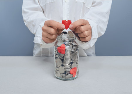 Woman in white shirt puts red heart in jar of silver coins. Love and romance concept. Love for moneyの写真素材