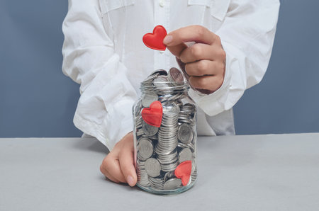 Donation jar with silver coins, into which the hand of a woman in a white shirt puts a red heart. Compassion conceptの写真素材