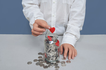 Donation jar with silver coins, into which the hand of a woman in a white shirt puts a red heart. Compassion conceptの写真素材