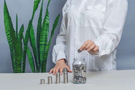 Woman in white shirt throws coins into glass jar, green plants. Concept of eco-friendly projects. Green businessの写真素材
