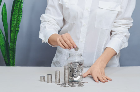 Woman in white shirt counting coins in glass jar, column of coins. Savings and financial planning conceptの写真素材