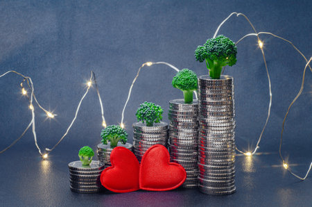 Two red hearts on columns of shiny coins with green trees illuminated by garlands. Dark blue background. Valentine's Dayの写真素材