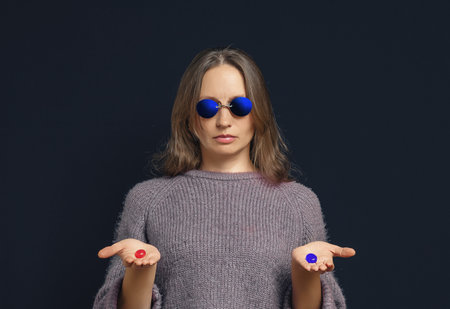 Serious woman in dark blue glasses holding blue and red pills on dark blue background. Choice conceptの写真素材
