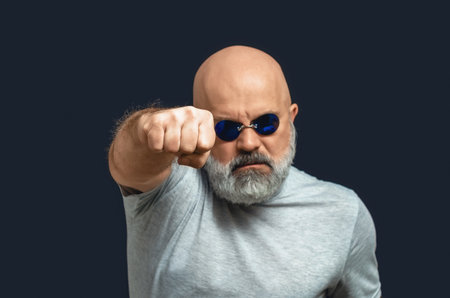 Bald man with gray beard in dark glasses looks at camera and seems to hit. Clenched fistの写真素材