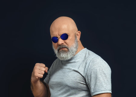 Bald man with gray beard in dark glasses looks at camera and raises his fist.の写真素材