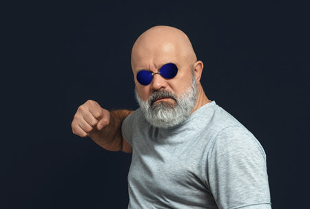 Bald man with gray beard in dark glasses looks at camera and raises his fist.の写真素材