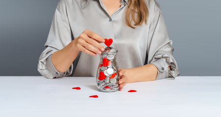 Donation jar with silver coins, into which the hand of a woman in a gray shirt puts a red heart. Compassion conceptの写真素材