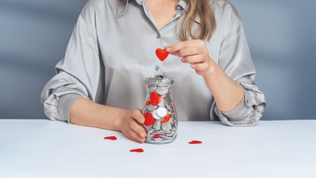 Donation jar with silver coins, into which the hand of a woman in a gray shirt puts a red heart....