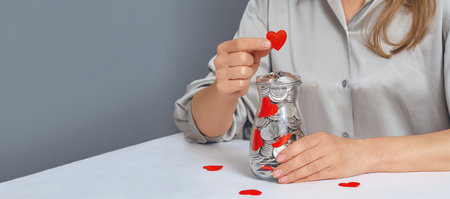 Donation jar with silver coins, into which the hand of a woman in a gray shirt puts a red heart. Compassion concept. Copy spaceの写真素材