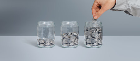 Three glass jars with coins as diagram. Female hand putting coin. Isolated on grey. Savings, finance, loans, pension conceptの写真素材