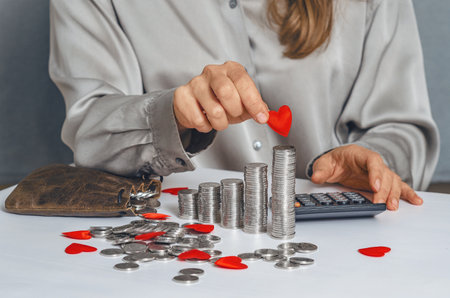 Woman in silver blouse holding small red heart over stacks of coins and counting on calculator....