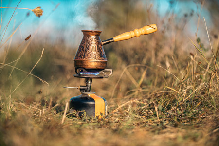 Coffee being prepared in copper engraved coffee pot on gas burner against blue blurred sky. Concept of relaxation and hiking in countryside on bikeの写真素材