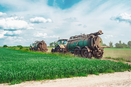 Side view of farm tractors with trailers containing tanks with organic fertilizers, manure. Tractors driving on road among green fieldsの写真素材