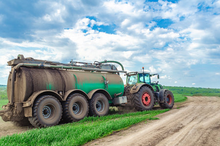 Tractor manure truck driving in field. Large tractor with trailer of liquid manure. Side view. White cumulus clouds in backgroundの写真素材