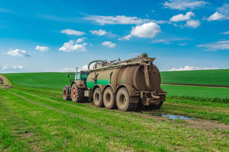 Tractor manure truck driving in field. Large tractor with trailer of liquid manure. Rear view. White cumulus clouds in backgroundの写真素材