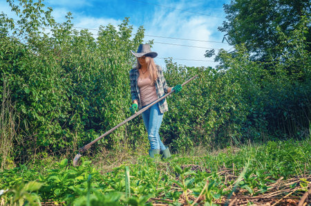 Smiling blonde woman in hat, plaid shirt, jeans and rubber boots mowing weeds with hand scythe on warm sunny day. Home yard care.の写真素材