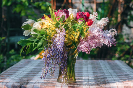 Vase with spring bouquet. Stylish spring bouquet on rustic table. Lilac, tulips, acacia and peonies in glass vase on checkered tablecloth. Floral still life.の写真素材