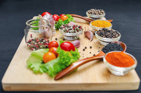 Wooden board with spices and vegetables. Cherry tomatoes, garlic, dry spices in bowls and spoons, on board on black background. Healthy food, cooking and vegetarian concept.の写真素材