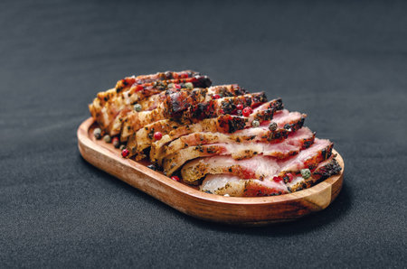 Fragrant slices of baked pork belly on a wooden board, sprinkled with colored peppercorns. Black...