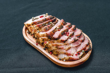 Fragrant slices of baked pork belly on a wooden board, sprinkled with colored peppercorns. Black background. Meat preparation concept.の写真素材